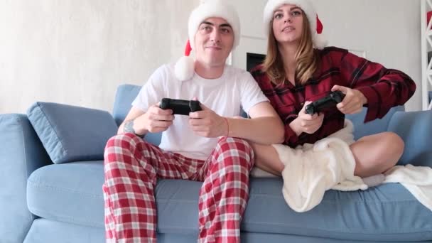Video games and holidays concept. Young blonde woman and caucasian man are holding joysticks sitting on sofa in living room, wearing santa hats, slow motion — Stock Video