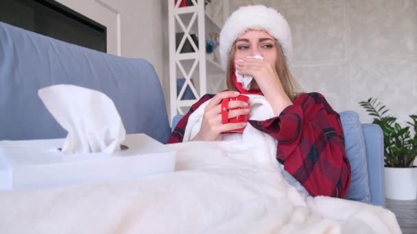 Κρύο ή ασθένεια έννοια. Νεαρή γυναίκα ξαπλωμένη σε καναπέ καλυμμένη με λευκό καρό κρατώντας ένα κόκκινο κύπελλο στο χέρι της φορώντας καπέλο Σάντα, αργή κίνηση — Αρχείο Βίντεο
