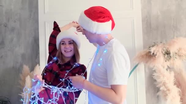 Holidays, love and relationships concept. Young woman and a man laughing and dabble, wrapped in a garland wearing santa hats, slow motion — Stock Video