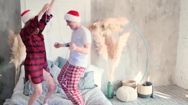 Boldog pár táncol az ágyon karácsonyi koszorúval otthon télapó kalapban és pizsamában — Stock videók