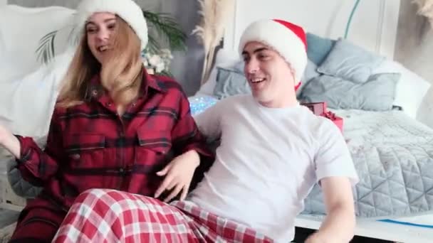 Fiatal boldog pár táncol és babrál a padlón az ágy mellett télapó kalapot és pizsamát visel, karácsonyi hangulat, lassított felvétel — Stock videók