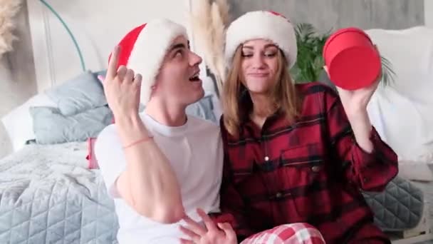 Glad man och kvinna dansar hemma i Santa hattar sitter på golvet nära sängen med gåvor på julafton bär pyjamas — Stockvideo