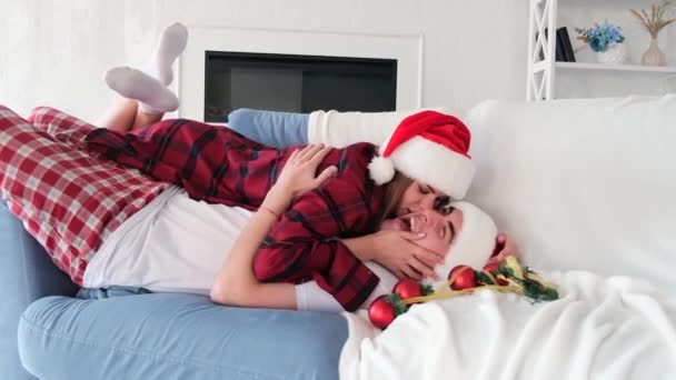 Jovem brincalhão encontra-se em cima do homem beijando-o e brincando no sofá cercado por decorações de árvore de Natal. Casal vestindo chapéus de Papai Noel e pijama xadrez vermelho — Vídeo de Stock