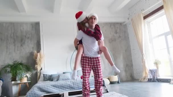 Julstämning. Unga par driver med varandra. Kvinna sitter ridande på baksidan av en man kramas och kysser honom klädd i tomte hatt och pyjamas — Stockvideo