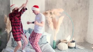 Happy couple dancing on the bed with Christmas garland at home wearing santa hats and pajamas, slow motion