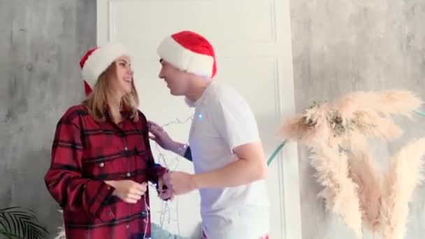 Casal feliz se divertindo com luzes de Natal em casa vestindo chapéus e pijamas — Vídeo de Stock