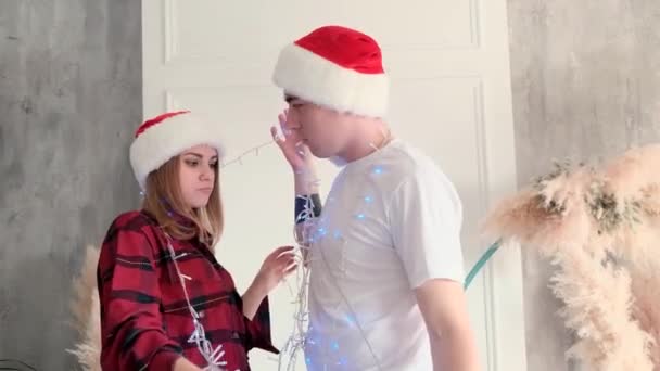 Férias, amor e relacionamentos conceito. Jovem mulher e um homem rindo e dabble, envolto em uma guirlanda usando chapéus de Papai Noel — Vídeo de Stock