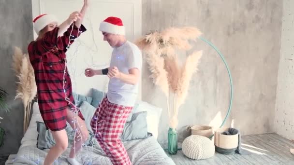 Casal feliz dançando na cama com guirlanda de Natal em casa vestindo chapéus e pijamas de Papai Noel, câmera lenta — Vídeo de Stock