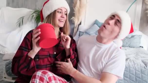 Young woman and man in Santa hats dance and have fun sitting on the floor near the bed on Christmas Day, slow motion — Stock Video