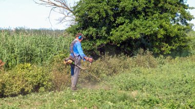 Kıdemli adam çimleri benzin fırçası makasıyla biçiyor. Tulumlu, koruyucu gözlüklü, ses geçirmez kulaklıklı ve iş eldivenli bir adam. Tam uzunluktaki kenar görünümü