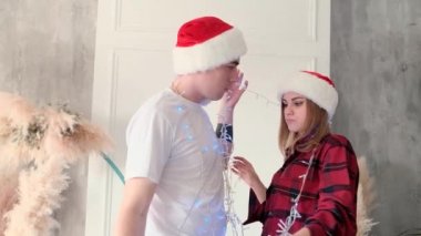 Mutlu çift evde Noel çelengiyle eğleniyor Noel Baba şapkası ve pijama giyiyorlar, ağır çekimde.