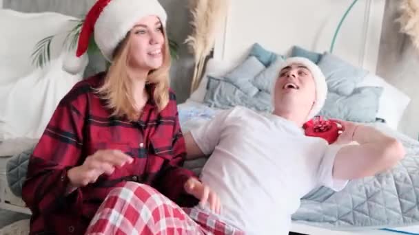 Jovem casal feliz dançando e dabbles no chão perto da cama vestindo chapéus de Papai Noel e pijama dando presentes para o Natal — Vídeo de Stock