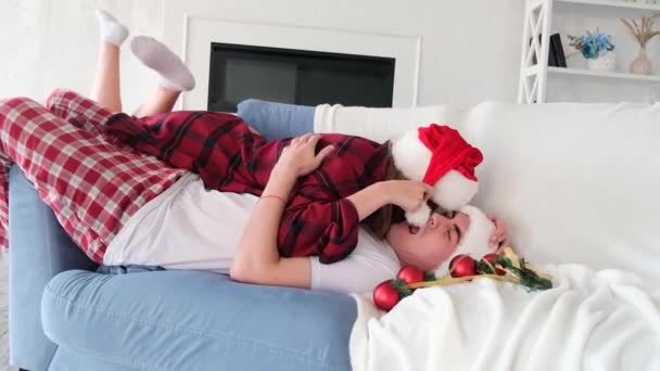 Playful young woman lies on top of man fooling around on the couch surrounded by Christmas tree decorations. Couple wearing santa hats and red plaid pajamas. Slow motion — Stock Video