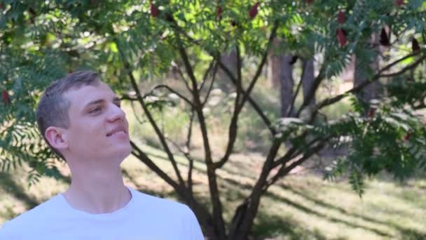 Portrait d'un jeune homme blond séduisant profitant de l'air frais dans le parc sur fond de buissons et d'arbres — Video