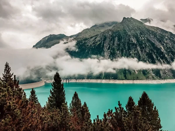 Veja através da floresta de abetos na água azul-turquesa da Bacia de Schlegeis, reservatório de montanha, envolto em nevoeiro e nuvens. Zillertal valley, Mayrhofen, Áustria — Fotografia de Stock
