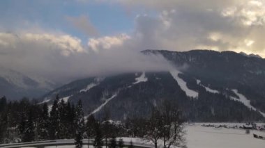Gün batımında orman ve bulutlarla kaplı karla kaplı dağların manzarası. Drone videosu