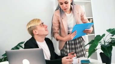 Ofiste iş akışı. Genç neşeli meslektaşlar bir şey hakkında tartışıyorlar. Sarışın adam masasında otururken, yanında kanekalon örgülü saçları olan modern kadın ellerinde klasörler tutuyordu..