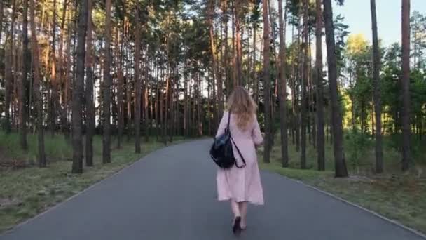 Vista trasera de la mujer rubia con una mochila negra caminando a lo largo de pinos altos en un camino de asfalto en un parque, la mujer lleva vestido largo rosa, 4k — Vídeo de stock