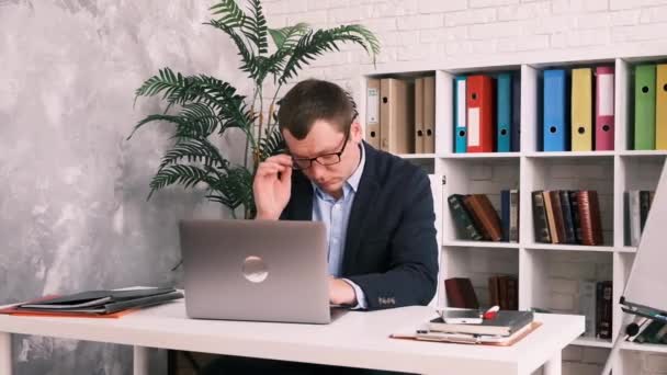 Szemfáradtság, rossz látás, irodai munka. Egy fiatalember leveszi a szemüvegét, és a kezével dörzsöli a szemét, miközben egy asztalnál ül egy laptoppal az öltönyében.. — Stock videók