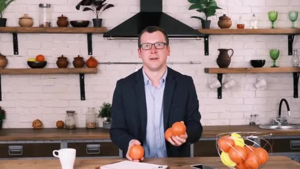 Jeune homme d'affaires joyeux dans des lunettes et un costume d'affaires jongle avec trois oranges tout en étant assis dans sa cuisine. Mouvement lent, pause du travail — Video