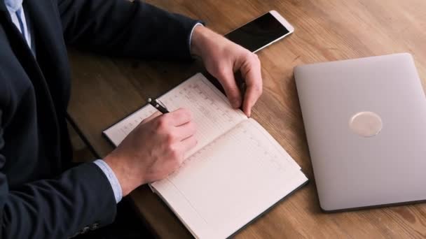 Vista superior de mãos masculinas preenchendo planejador ou bloco de notas ao lado de laptop e telefone celular na mesa de madeira. Conceito de negócios, planejamento e gerenciamento de tempo. Um homem de fato. Colarinho branco trabalhador — Vídeo de Stock