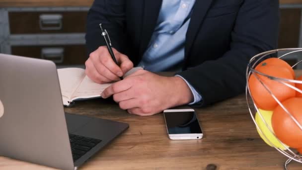 Vista ritagliata delle mani maschili che compilano il pianificatore o il blocco note accanto al computer portatile aperto e al telefono cellulare sul tavolo di legno. Business, pianificazione e gestione del tempo. L'uomo indossa un completo. Lavoratore colletto bianco — Video Stock