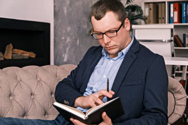 Takım elbiseli ve gözlüklü başarılı genç adam şöminenin arka planında geniş bir ofiste otururken defter ya da planlamacıyla çalışıyor. Başarı, iş ve zaman yönetimi kavramı