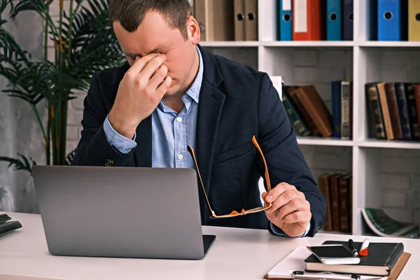 Szemfáradtság, rossz látás, irodai munka. Egy fiatalember leveszi a szemüvegét, és a kezével dörzsöli a szemét, miközben egy asztalnál ül egy laptoppal az öltönyében. Lassú mozgás. Stock Fotó