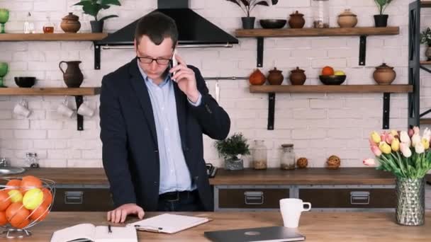 Naštvaný obchodník v brýlích a obleku hádky s někým na telefonu, zatímco stojí v kuchyni. Křičí do telefonu, bouchne pěstí do stolu, odhodí telefon na stůl a odejde. — Stock video