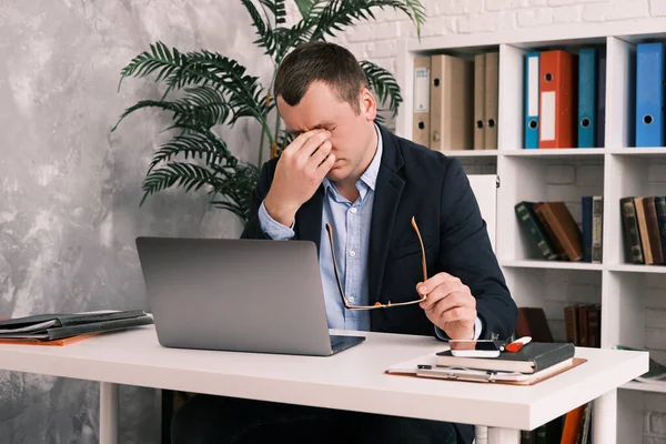 Eye fatigue, poor eyesight, office work. A young man takes off his glasses and rubs his eyes with his hand while sitting at a table with a laptop in a suit. Slow motion — Stock Photo, Image