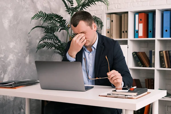 Ögontrötthet, dålig syn, kontorsarbete. En ung man tar av sig glasögonen och gnider blicken med handen när han sitter vid ett bord med en laptop i kostym. Långsamma rörelser Royaltyfria Stockfoton