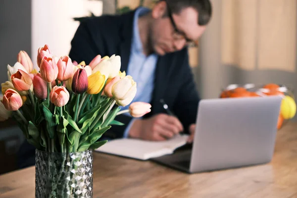 Bouquet of tulips in the foreground. Businessman wearing suit and glasses is writing something in a notebook sitting behind a laptop in the background. Selective focus Стоковое Изображение