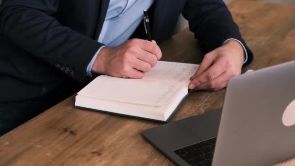 Vue latérale de l'homme d'affaires en costume remplissant planificateur ou bloc-notes à côté d'un ordinateur portable ouvert contre de bouquet frais de tulipes. Un gestionnaire ou un cols blancs pratique la gestion du temps. Mouvement lent — Video