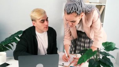 Ofiste projeyi tartışan iki kadın ve erkeğin süper yavaş çekimleri. Ceketli adam sandalyede oturuyor, uzun ekose etekli kadın ve ceket onun yanında duruyor.