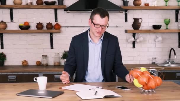 Relaxed male entrepreneur in business suit and glasses throws an orange from hand to hand while sitting at home in kitchen, looking at notebook lying on the table, pondering information. Slow motion. — Stock Video