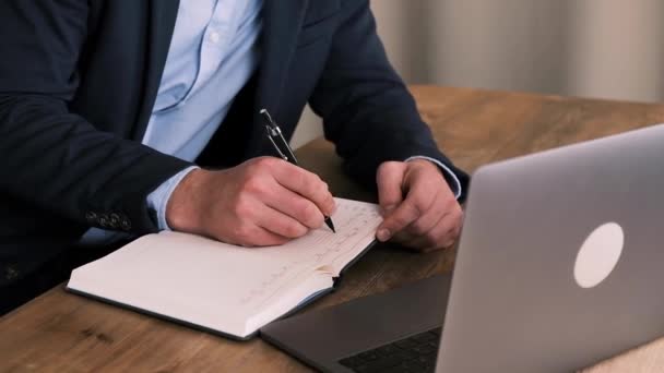 Vista lateral recortada del hombre de negocios en traje escribiendo algo en el cuaderno con una computadora portátil abierta en primer plano. En tiempo real. Gerente o trabajador de cuello blanco llena el planificador — Vídeos de Stock