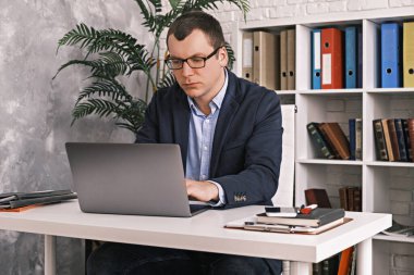 Ciddi, odaklanmış, gözlüklü ve takım elbiseli bir adam dizüstü bilgisayarla bir şeyler yazarken arka planda klasörlerle birlikte açık bir ofiste oturuyor. İş ve girişimcilik kavramı