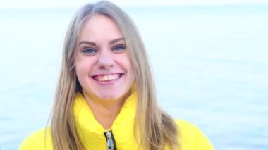 Genç, mutlu, mavi gözlü bir kadın güneşli bir bahar gününde sarı bir ceketle nehrin arka planına bakıyor. Hayatın zevki, olumlu tutum. Yavaş çekim, 4k