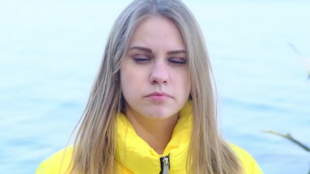 Портрет грустной серьезной молодой женщины, смотрящей в камеру на фоне реки в ярко-желтой куртке весной или осенью солнечного дня. Размышления, разочарование, тревога — стоковое видео