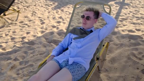 Joven empresario divertido en una camisa, corbata, ropa interior y gafas de sol se encuentra en la playa en la tumbona y disfruta de unas vacaciones de verano, lanzando su mano detrás de la cabeza, cámara lenta, 4k — Vídeos de Stock