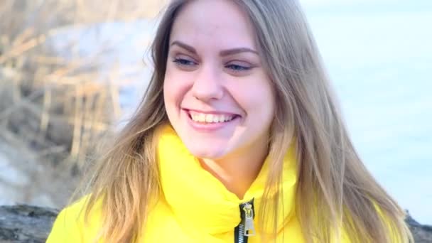 Молодая счастливая весёлая улыбающаяся застенчивая голубоглазуя женщина смотрит в камеру на фоне природы в желтой куртке в солнечный весенний день. Внутренняя гармония, умиротворение. 4k, реальное время — стоковое видео