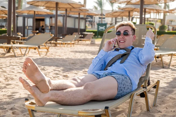 Giovane uomo d'affari felice downshifter parla al telefono sdraiato su un lettino in mezzo alla spiaggia in camicia, cravatta e boxer — Foto Stock