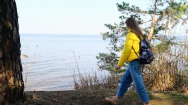 Sırt çantalı dişi turist, büyük bir göl veya nehrin kıyısındaki ormanda yürüyüş yapmakla meşgul. Güneşli bir bahar gününde göletin kenarına yürür ve manzaranın tadını çıkarır. 4k, ağır çekim.
