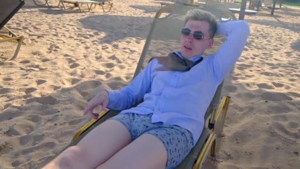 Ung affärsman i skjorta, slips, underkläder och solglasögon ligger på stranden på solstolar. Hör samtal, lyfter luren och lägger den åt sidan, knäpper upp skjortan och kliar sig i magen. Långsamma rörelser, 4k — Stockvideo