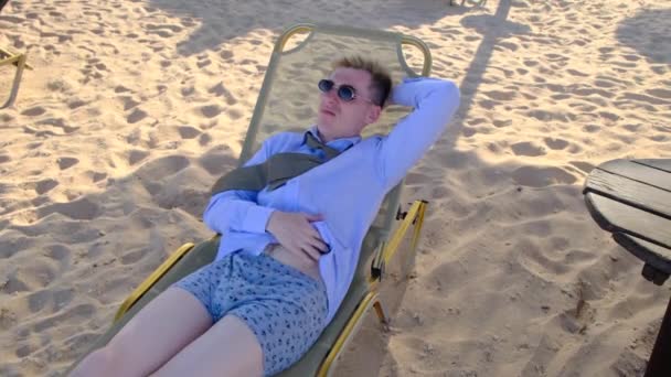 Businessman in shirt, tie, underwear and sunglasses scratching his belly, lying on beach on sun lounger and enjoying summer holidays, throwing his hand behind his head, slow motion, 4k — Stock Video