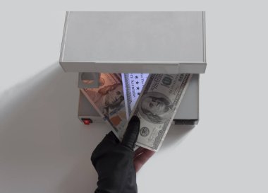 Siyah eldivenli bir el doğrulama için üç banknot tutar. Para birimi otantik detektörünün ultraviyole lambası sahte banknotu parlak bir şekilde aydınlatıyor. Gerçek banknotlar parlamaz.