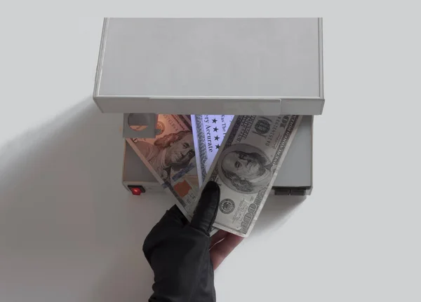Siyah eldivenli bir el doğrulama için üç banknot tutar. Para birimi otantik detektörünün ultraviyole lambası sahte banknotu parlak bir şekilde aydınlatıyor. Gerçek banknotlar parlamaz.