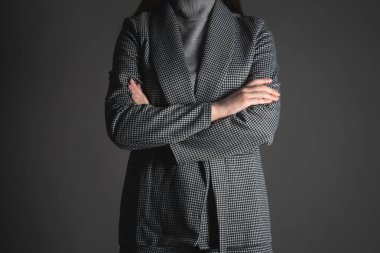 Gri arka planda elleri çapraz duran takım elbiseli bir kadın var..