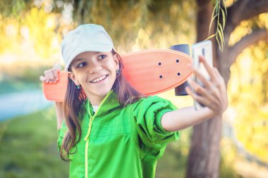 Küçük mutlu hippi kız elinde kaykayla cep telefonuyla selfie çekiyor.