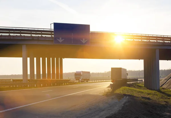 Trucks with semi-trailers move along the highway against the backdrop of a sunny sunset. Automobile tie and bridge on the background of the sun. Logistics and cargo transportation concept, commerce